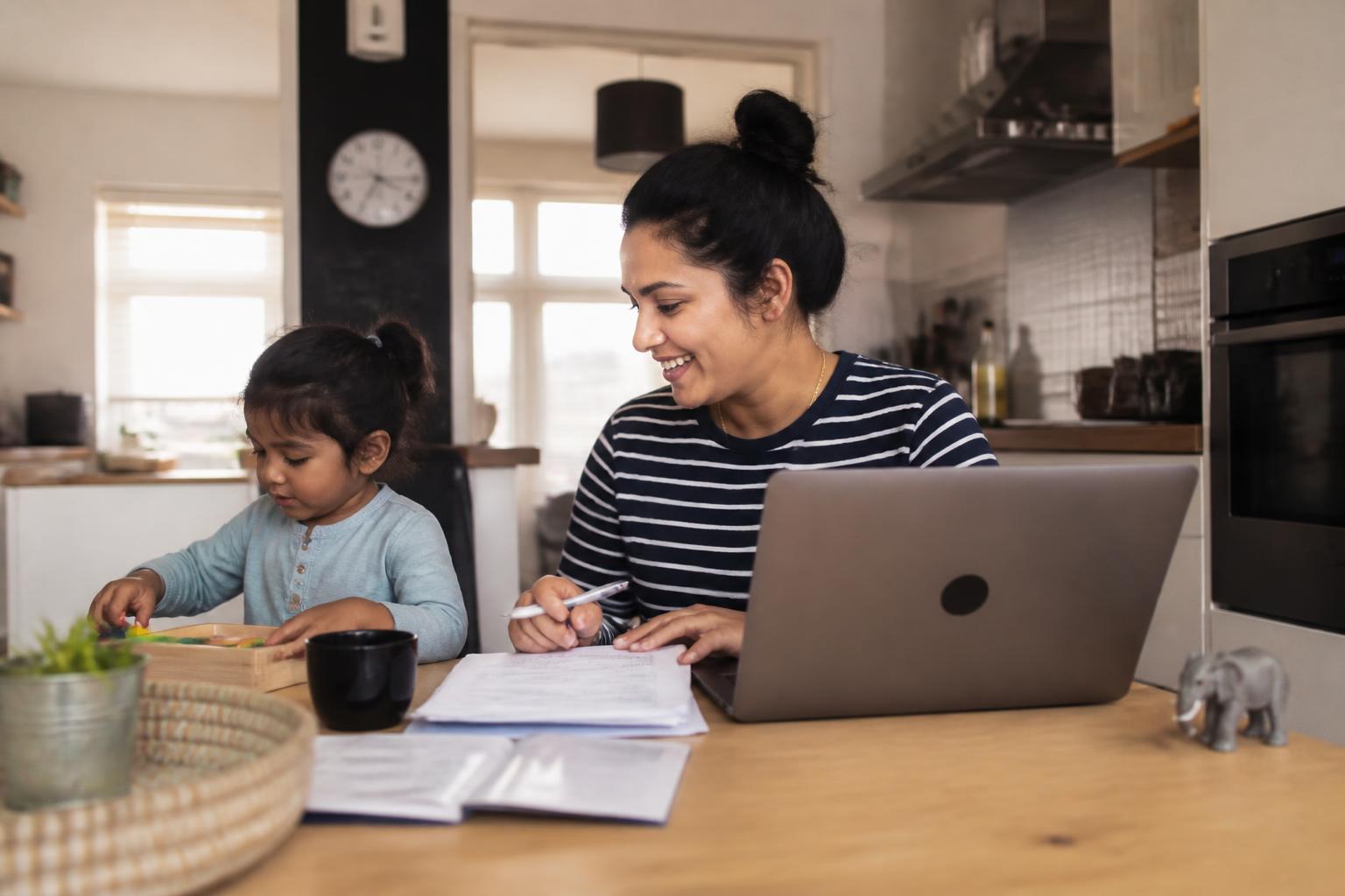 Moeder regelt de kinderopvangtoeslag op haar laptop terwijl haar dochter naast haar speelt